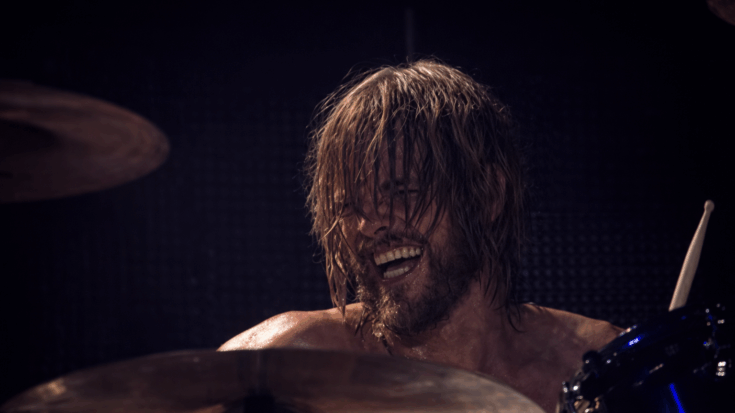 Taylor Hawkins plays the drums with a wide smile, his sweat-soaked hair flying as he powers through a high-energy performance.