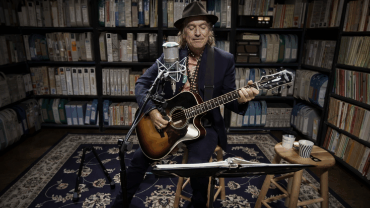 Dave Davies plays an acoustic guitar in a cozy room lined with shelves, performing into a studio microphone.