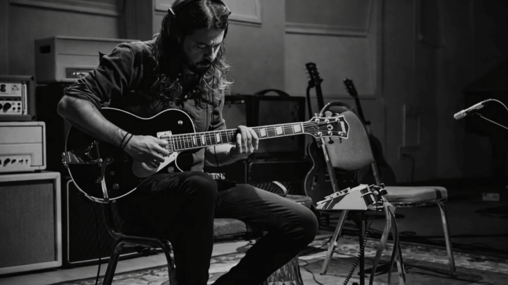 Dave Grohl sits focused in a studio, playing an electric guitar during a quiet recording session.