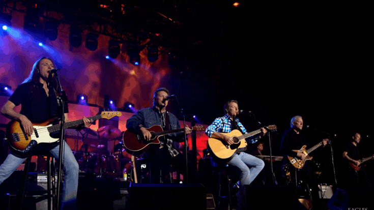 The Eagles perform under glowing stage lights, with acoustic guitars at center, electric guitars on the sides, and a bass driving the left side of the stage.