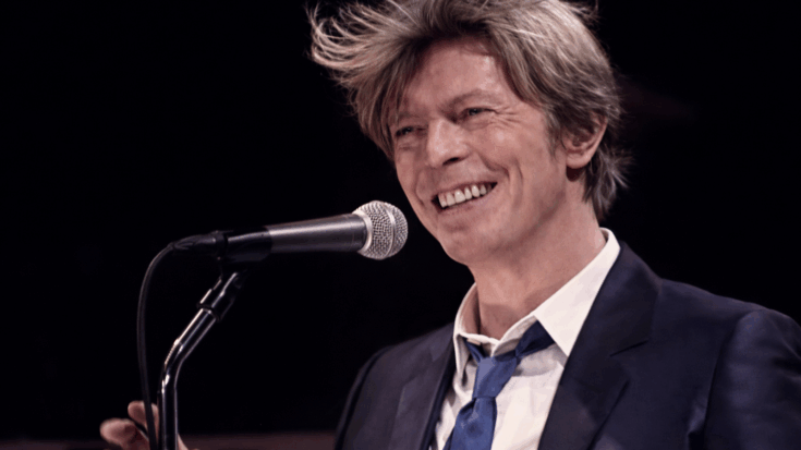 David Bowie smiles at the microphone in a sharp suit, with his windswept hair adding to the moment’s charm.