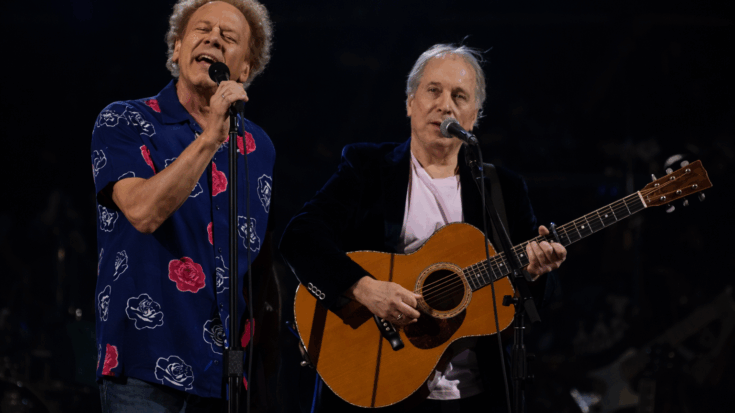 Simon and Garfunkel perform together onstage, with Art Garfunkel singing and Paul Simon playing an acoustic guitar.