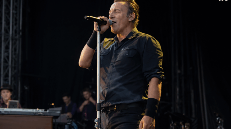Bruce Springsteen delivers a powerful live performance at the microphone in a dark stage setting.