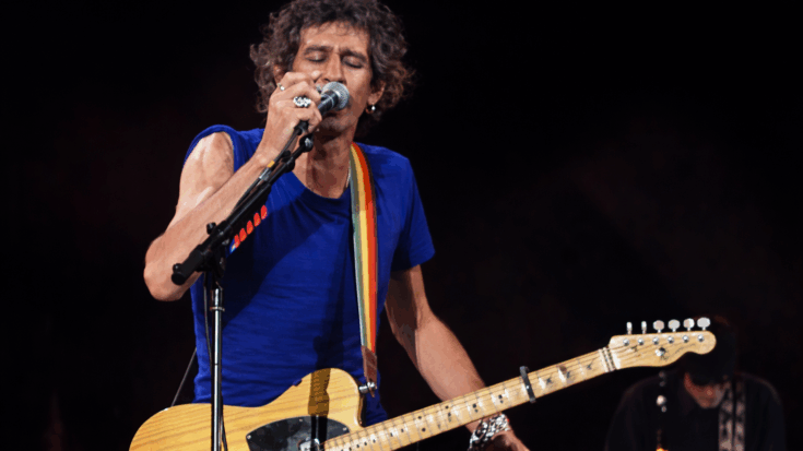 Mick Jagger sings into the microphone while playing guitar onstage in a blue shirt.