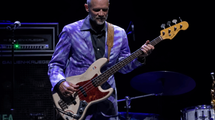Flea performs onstage with his bass while wearing a striking purple suit under dark concert lighting.