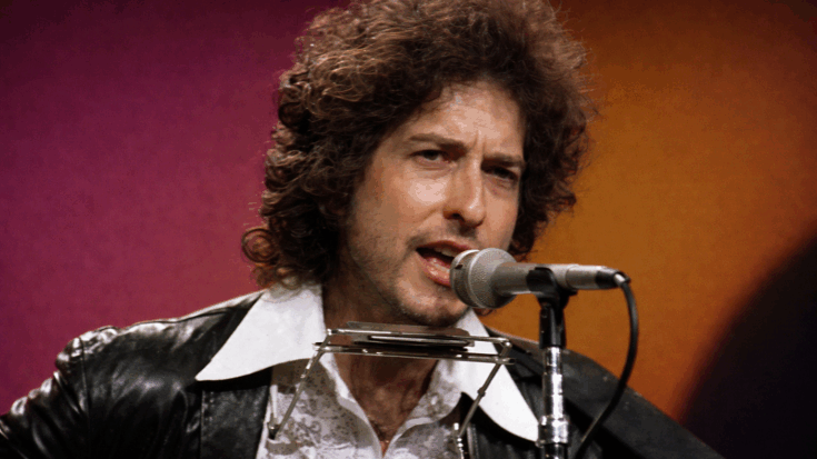 Bob Dylan sings into a microphone with a harmonica holder around his neck during a live performance.