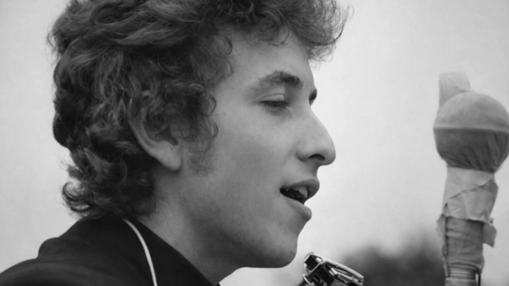 Bob Dylan sings in a black-and-white close-up with a harmonica holder around his neck.