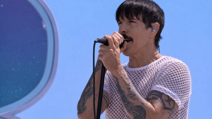 Anthony Kiedis sings into a microphone outdoors while wearing a white mesh shirt.