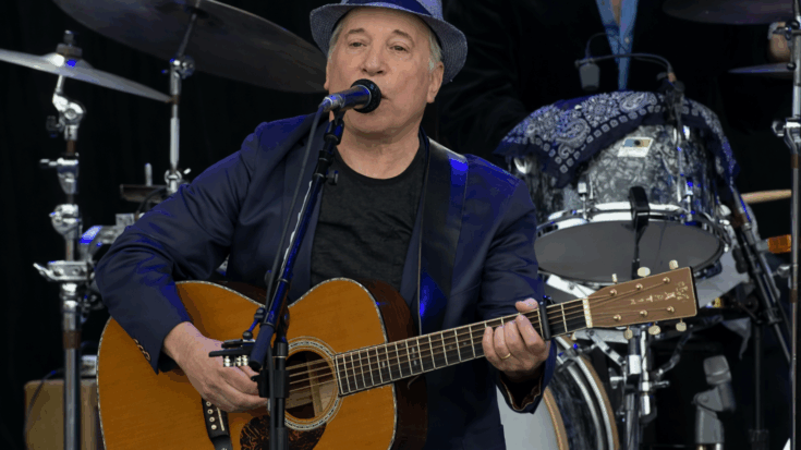 Paul Simon performs onstage with an acoustic guitar while singing into the microphone.