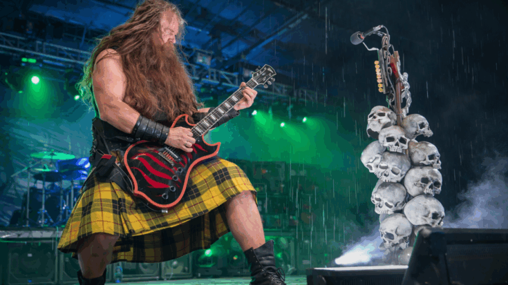 Zakk Wylde performs powerfully onstage in the rain while playing a red-and-black bullseye-style electric guitar.