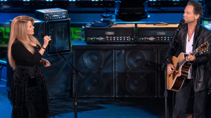 Stevie Nicks plays an acoustic guitar while Lindsey Buckingham sings beside her during a live duet onstage.