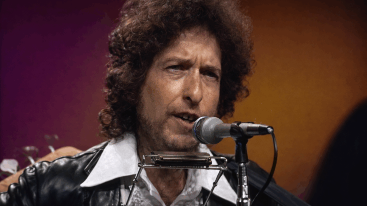 Bob Dylan sings into a microphone with a harmonica holder around his neck during a live performance.