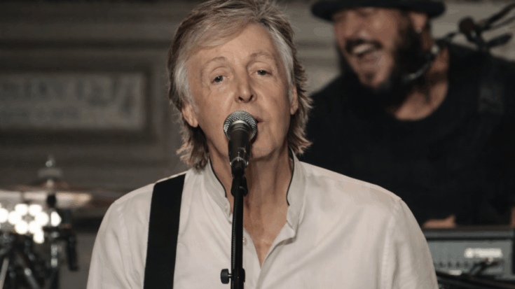 Paul McCartney sings into a microphone during a live performance while wearing a white shirt.
