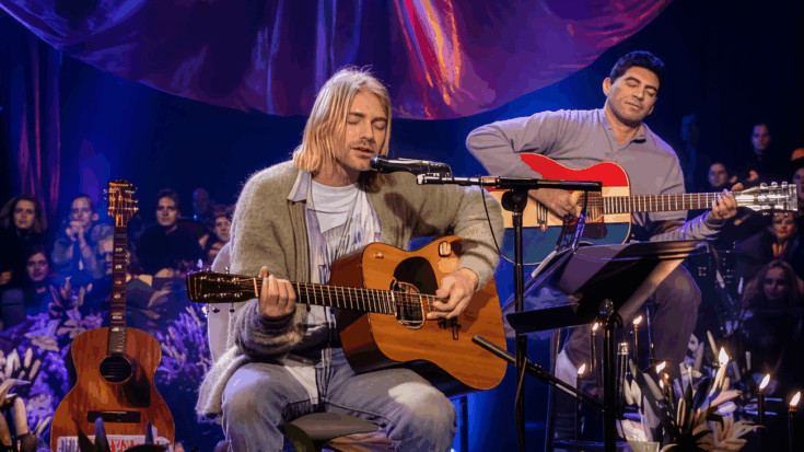 Kurt Cobain sits onstage, softly singing while strumming an acoustic guitar in an intimate, candlelit performance.