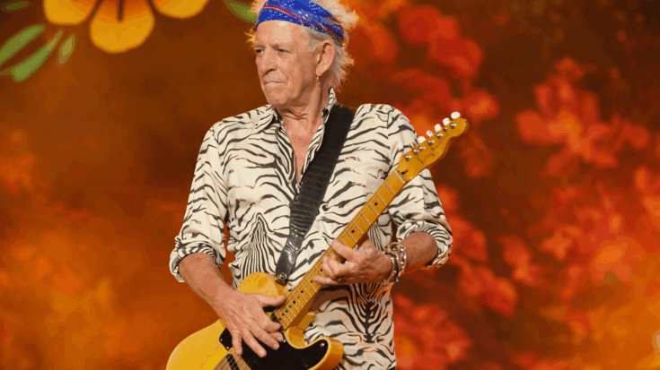 Keith Richards plays a yellow electric guitar on stage, wearing a zebra-print shirt and a blue bandana against a warm, vibrant backdrop.