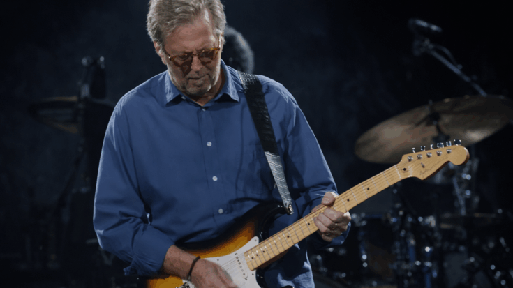 Eric Clapton looks down while playing a sunburst electric guitar, wearing glasses and a blue button-up shirt on stage.