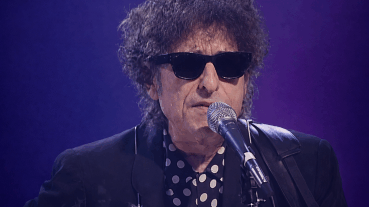 Bob Dylan performs on stage wearing dark sunglasses and a polka-dot shirt, singing into a microphone with a harmonica holder around his neck.