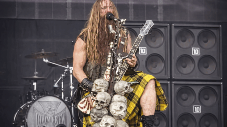 Zakk Wylde performs on stage with his signature long hair and beard, playing a skull-adorned guitar while singing into a microphone.