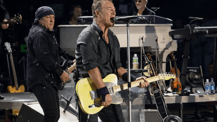 Bruce Springsteen passionately sings into a microphone while strumming a bright yellow electric guitar during a high-energy live performance on stage.