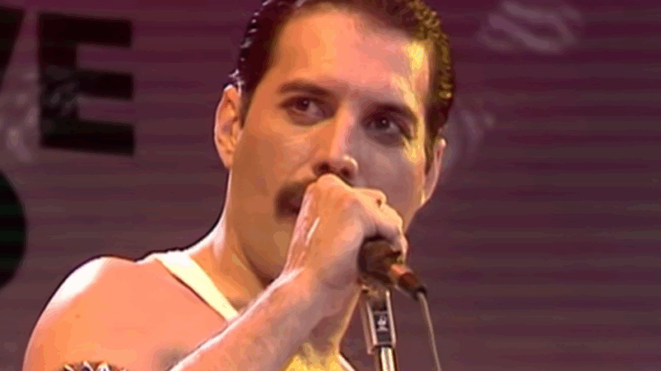Freddie Mercury commands the stage in a white tank top, his powerful voice cutting through the red-lit atmosphere with electrifying intensity.