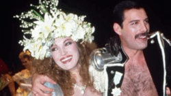 Jane Seymour and Freddie Mercury smile together in glamorous costumes, with Mercury playfully posing while Seymour wears an elaborate floral headpiece.