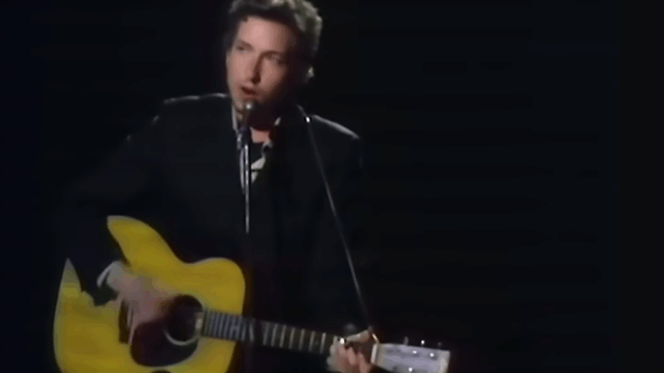 Bob Dylan gently commands the stage, cradling his steel-string Martin D-28–style dreadnought as he delivers a calm, soulful performance.