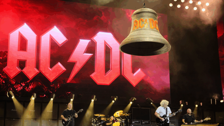 AC/DC performs live onstage beneath a giant glowing logo and a hanging bell, with the band rocking under bright concert lights.