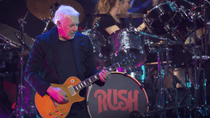 Rush guitarist Alex Lifeson performs with focused intensity on a brightly lit stage while a drummer plays behind him amid colorful concert lights.