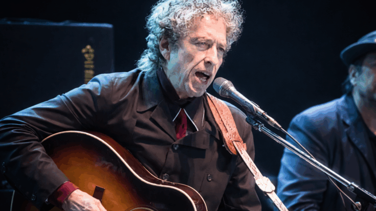 Bob Dylan sings into a microphone while playing an acoustic guitar under cool stage lights.