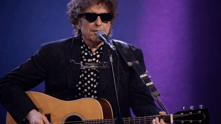 Bob Dylan is singing into a microphone while playing an acoustic guitar, wearing sunglasses under soft stage lighting.