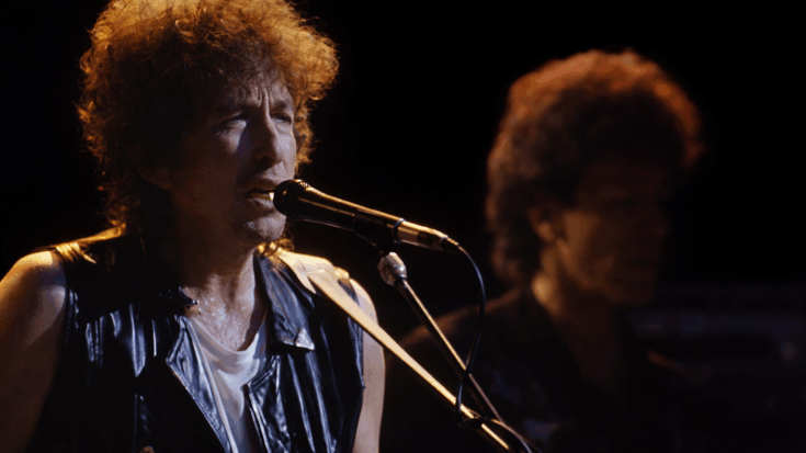 Bob Dylan is singing into a microphone onstage while another musician stands blurred in the background.