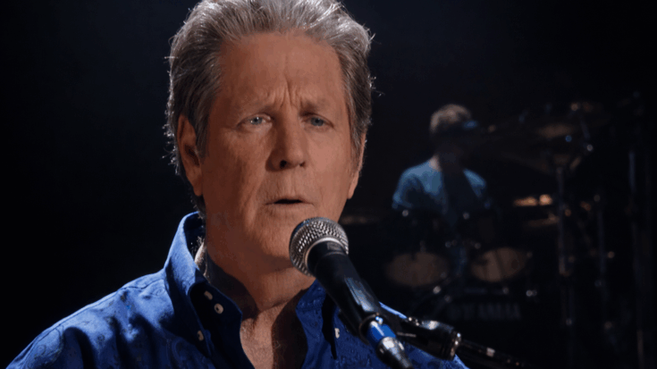 Brian Wilson sings into a microphone with a calm, focused expression under soft stage lighting.