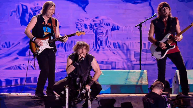 Iron Maiden performs live with the singer crouched at center while two guitarists play under vivid blue and purple stage lights.