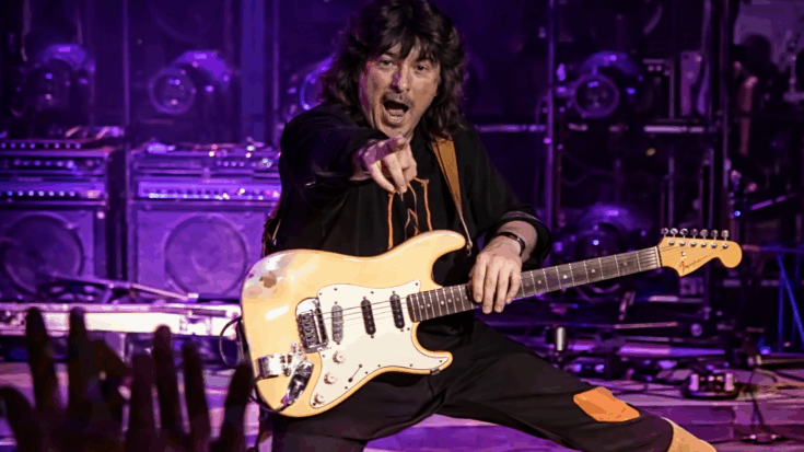 Ritchie Blackmore points toward the crowd while holding a white guitar under purple stage lights.