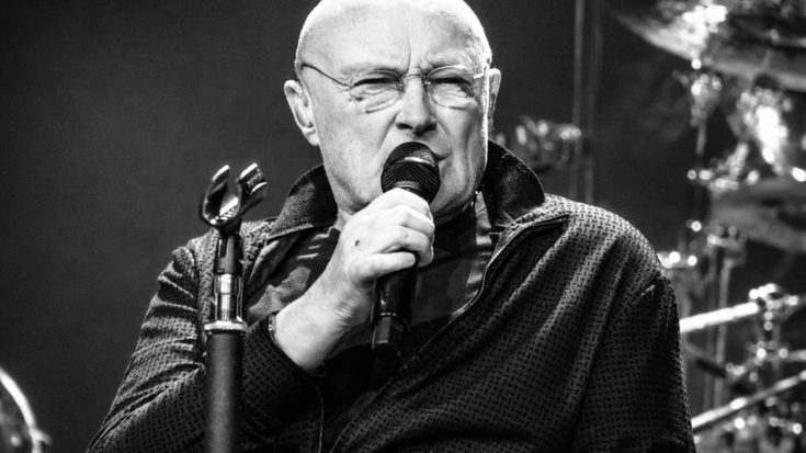 Phil Collins sings into a microphone on stage, captured in a close-up with a serious, focused expression.