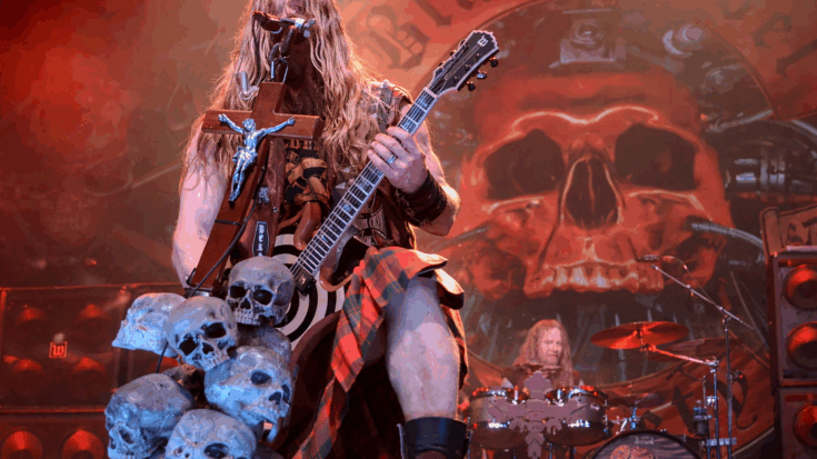 Zakk Wylde performs on stage with his bullseye guitar, surrounded by skull props and a fiery backdrop.