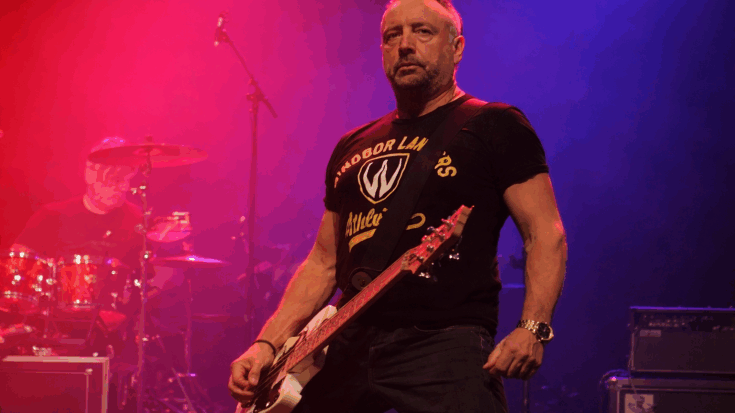 Peter Hook stands on stage holding his bass, lit by red and blue lights with a focused, intense expression.