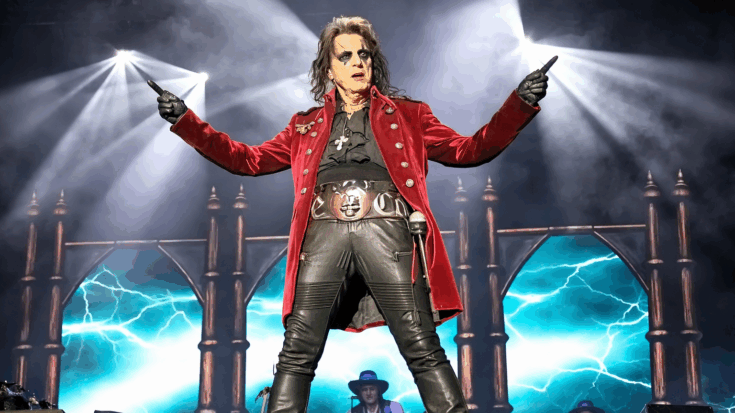 Alice Cooper stands on stage in a red military-style coat and black leather pants, striking a dramatic pose under bright concert lights.