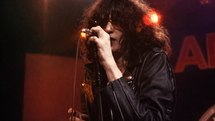 Joey Ramone grips the microphone in a raw live performance, wearing a black leather jacket with his signature shaggy hair partly covering his face.