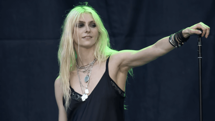 Taylor Momsen stands on stage in a black outfit, holding a microphone stand with a calm, confident expression under green lighting.