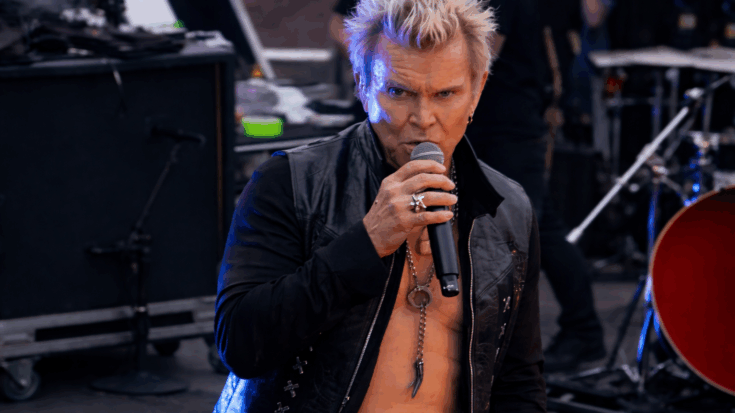 Billy Idol grips a microphone mid-performance, showing his signature snarl and spiky blonde hair in a high-energy moment.