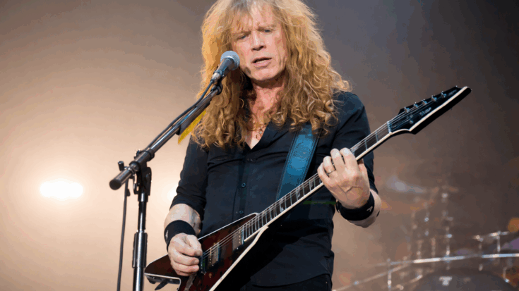 Dave Mustaine plays his guitar while singing into a microphone, his long curly hair framing an intense onstage expression.