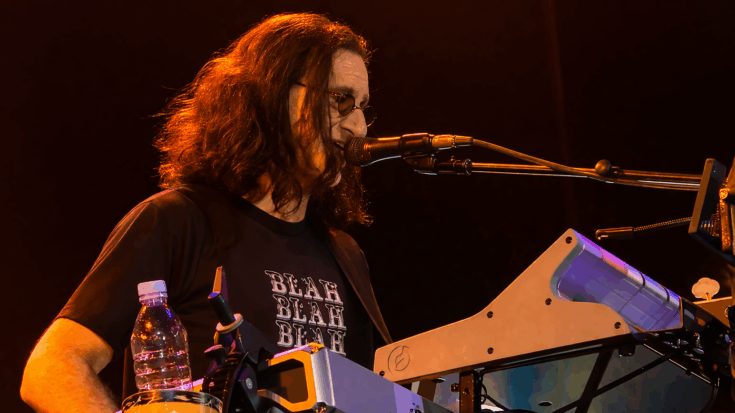 Geddy Lee performs at his keyboard, singing into a microphone under warm stage lighting with his signature long hair and glasses.
