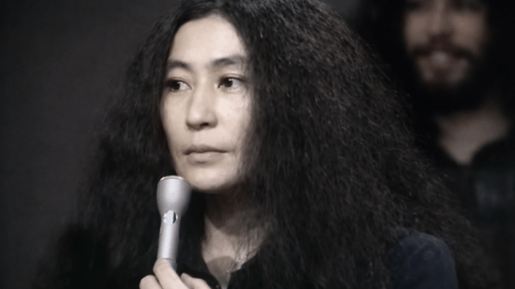 Yoko Ono holds a microphone with a calm, focused expression, her long textured hair framing her face.