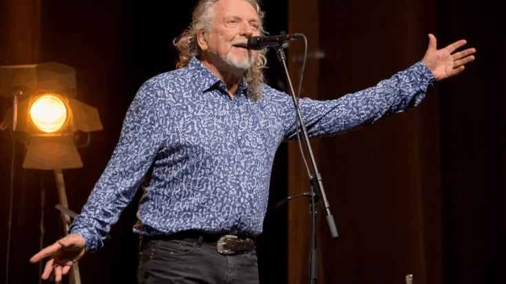 Robert Plant stands at the microphone with arms outstretched, singing under warm stage lighting.