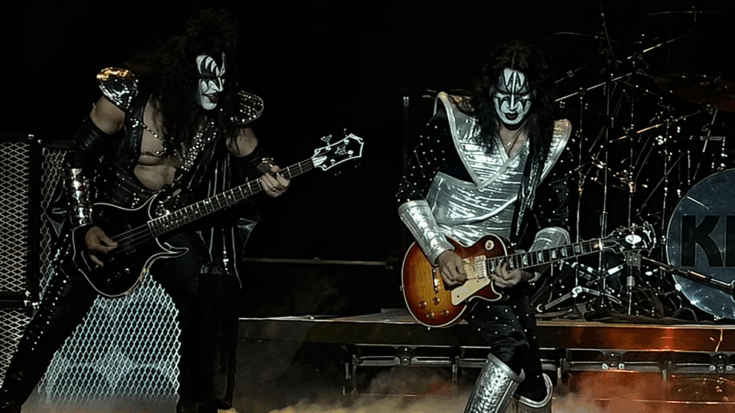 KISS performs onstage in full makeup and metallic costumes, playing guitars through smoke and dramatic concert lighting.