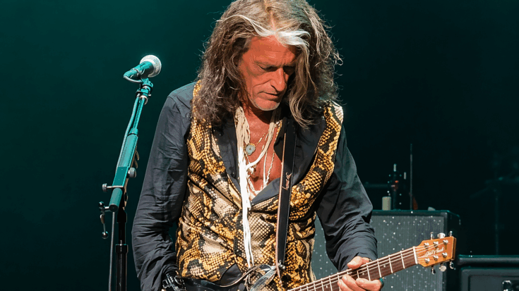 Joe Perry looks down at his guitar while performing onstage, wearing a patterned vest and long loose hair under soft green lighting.