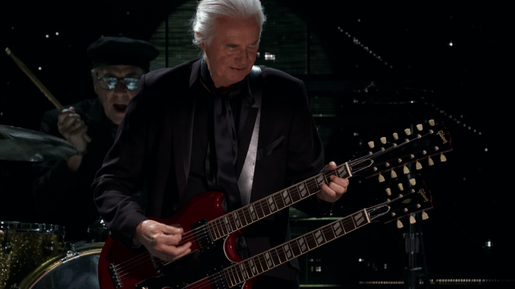 Jimmy Page plays a red double-neck guitar onstage, dressed in black under dark concert lighting.
