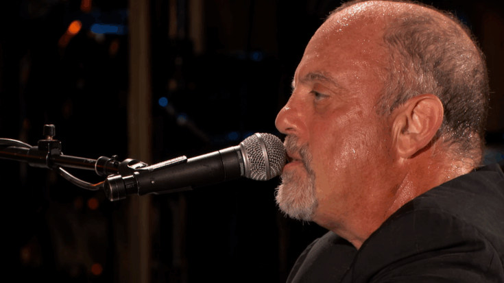 Billy Joel sings into a microphone in a close-up performance shot, his face glistening under warm stage lights.