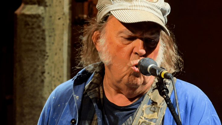 Neil Young sings into a microphone outdoors, wearing a cap and layered casual clothing in a candid performance moment.
